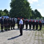 DSC_0033 Na obrazku widnieje grupa umundurowanych osób, z których korzystają na głowach czerwonych czapki; wszystkie rozwiązania w szeregu na tle przyrody. W widocznych drzewach oraz ludzi ubranych w codziennych strojach.