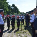 DSC_0035 Na obrazku grupa umundurowanych osób stoi w formacji na świeżym powietrzu, zwrócona w stronę kilku oficjeli w odświętnych strojach. W tle można spędzić kilku obserwatorów oraz namiot, a wszystko na miejscu w słoneczny dzień.