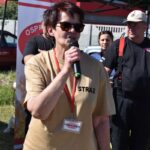 DSC_0045 Na obrazie widać osobę w okularach graficznych i beżowej koszuli z napisem „STRAŻ”, która przemawia do mikrofonu podczas wydarzeń plenerowych. W tle znajdują się dwie inne osoby, z których jedna jest ubrana w czarnym mundurze i kapeluszu.