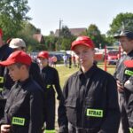 DSC_0054 Zdjęcie przedstawia grupę ludzi w czarnych mundurach z naszymiywkami „STRAŻ” oraz czerwonemi czapkami, stojącymi na świeżym powietrzu w słoneczny dzień. W tle widoczne są różne drzewa i akcesoria.