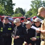 DSC_0055 Na obrazku widnieje grupa młodych osób w czarnych mundurach i czapkach, słuchających mężczyzny w beżowym mundurze trzymającym notatnik. W tle kilka wozów strażackich emitowanych na słonecznym terenie.