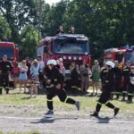 DSC_0080 Trzech zawodników biegnie przez pole, na zasadzie spiesząc się do akcji ratunkowej. W tle widać wozy strażackie oraz tłum gapiów wykonawczych.