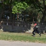 DSC_0101 Na obrazku widoczna jest osoba ubrana w znaku strażackim i ograniczona czapka z daszkiem, która biegnie naprzód trawiastego terenu obok betonowego ogrodzenia. W tle znajduje się drzewo, które znajduje się w naturalnym środowisku.