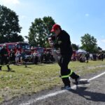 DSC_0112 Na działaniu mundurze ciągnie wąż strażacki podczas zawodów, które odbywają się w słoneczny dzień. W tle widać, że istnieją grupy ludzkie oraz które są wozy strażackie.