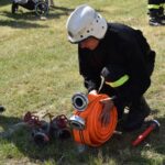 DSC_0117 Na obrazku wodnym w ekwipunku kuca na trawie, zwijając pomarańczowy wąż obok kilku metalowych zaworów. W tle widoczni są ludzie i wózki dziecięce.