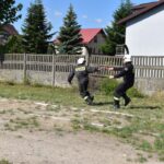 DSC_0123 Na obrazku dwaj strażacy w mundurach biegnących obok ogrodzenia, z jednym szybko przychodzącym drugim. W tle widoczne są domy i drzewa.