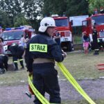 DSC_0128 Na obrazku widocznym w ochronnym ubraniu z napisem „STRAŻ”, który trzyma żółty wąż pożarniczy. W tle znajdują się wozy strażackie oraz ludzie, którzy są częścią akcji.