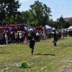 DSC_0133 Na obrazku dwie osoby w ochronnym stroju biegnącym przez trawiastą polanę, a w tle widoczne są wozy strażackie oraz zgromadzeni obserwatorzy. Wydaje się, że scena stanowi akcję ratunkową lub działalność produkcyjną.