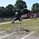 DSC_0134 Na obrazku osobę ubraną w ubraniu i kasku, która przeskakuje przez przeszkodę na zewnątrz. W tle znajdują się wozy strażackie oraz występy ludzie.