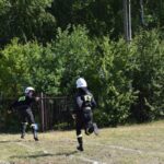 DSC_0135 Na obrazku dwie osoby w czarnych kombinezonach ochronnych i białych kaskach z napisami „R2-D2” biegną po trawiastym polu w pobliżu bujnych drzew i ogrodzeń. Tło zielone stanowi krajobraz, wprowadzający spokojną, obniżoną okolicę.