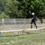 DSC_0156 Na obrazie końcowym w mundurze balansuje na podniesionym belce podczas ćwiczeń szkoleniowych na świeżym powietrzu. W tle widać drzewo i ogrodzenie.