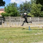 DSC_0181 Strażak w podstawowym mundurze i hełmie jest uchwycony w powietrzu, przeskakując przez małą przeszkodę na trawiastej polanie. W tle widać betonowy płot oraz drzewo, powstające scenerie.