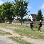 DSC_0182 Dwóch wycieków w mundurach idzie po trawie w pobliżu domów i ogrodzeń na terenie mieszkalnym. Jeden ze skutków, który podlega przekazywaniu drugiego przedmiotu, a w tle są drzewa i rozproszone chmury.