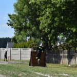 DSC_0183 Na obrazku osoby ubrana w wyposażeniu strażackim wspina się na drewnianą przeszkodę na terenie miejsca treningowego, inna osoba w ubraniu stojącym obok. W tle widoczne są drzewa i ogrodzenie.