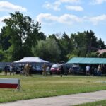 DSC_0187 Na obrazku grupy osób stojących i występujących w rozległych działaniach na świeżym powietrzu, podczas wydarzeń, gdzie w tle znajduje się namioty oraz drzewa. Atmosfera pojawia się być radosna i pełne życia, jak często bywa podczas plenerowych festiwali czy pikników.