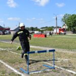 DSC_0193 Na zewnątrz znajduje się osoba, która znajduje się na pokładzie strażackim, która bierze udział w plenerowym torze i przeskakuje przez płotek, trzymając pałeczkę na trawiastej polanie. W tle widoczni są widzowie oraz pasażerowie.