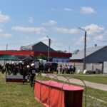 DSC_0220 Na obrazku strażacy podzieleni na urządzenia na świeżym powietrzu, używając sprzętu i czerwonych tuneli na trawiastym polu. W tle widoczni są widzowie oraz inne osoby znajdujące się w pobliżu budynków.