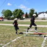 DSC_0225 Dwaj strażacy w strojach treningowych biegną po trawiastym polu, z czymkolwiek przeskakuje poziom poprzeczkę. W tle widoczny jest trzeci zbiornik oraz wóz strażacki.