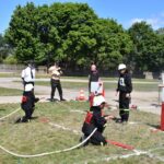 DSC_0232 Na obrazku ludzi w mundurach strażackich, wyodrębniających udział w działaniu przeciwpożarowym na świeżym powietrzu, używających węży niszczyzych oraz celów. W tle znajduje się drzewo i dodatkowi uczestnicy szkoleń.