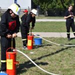 DSC_0238 Na obrazku dzieci ubrane w strażackie mundury obsługujące pompy wodne na świeżym powietrzu. W tle widać inne dzieci oraz osobę dorosłą, którzy przypatrują się wydarzeniu.