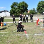 DSC_0245 Na obrazku strażacy uczestniczyli w ćwiczeniach na świeżym powietrzu, używając węży do celowania w panelach. Grupa obserwatorów, monitorowane w mundurach, obserwujące działania i notatki.