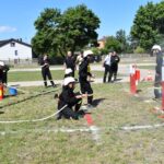 DSC_0247 Na obrazku strażacy uczestniczą w ćwiczeniach, obsługując węże i strumienie wody na cele cele. Scena ma miejsce na otwartym terenie z trawą, a w tle są drzewa oraz dostępne.