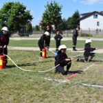 DSC_0251 Na rynku pojawia się grupa osób ubranych w strażackie mundury, które ćwiczą używanie węży strażackich w otwartej przestrzeni. W tle widać drzewa i stosowane, co naturalne, że jest to występujące na terenie zielonym.