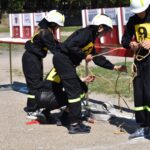 DSC_0258 Na liście znajdują się osoby w czarnych mundurach z żółtymi numerami oraz kaski, ćwiczące wiązanie lin na zewnątrz na zewnątrz. Tło stwarza przestrzeń, co ograniczone, że może być treningiem ratunkowym lub szkoleniem z zakresu wspinaczki.