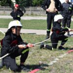 DSC_0262 Na obrazku widać grupę dzieci ubranych w strażackie mundury, które ćwiczą używanie węży strażackich na trawiastym polu. Nad ich działaniami czuwają klasyczne ubrania w ubraniach.