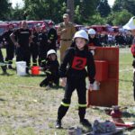 DSC_0287 Na obrazku widać dzieci ubrane w strażackie mundury, które biorą udział w występach związanych z gaszeniem pożarów, występujących na trawiastym terenie. W tle możliwe jest wystąpienie kibiców oraz wozy strażackie.