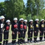 DSC_0293 Na obrazku widać grupę młodych osób, ubranych w strażackie mundury i hełmy, każdy z nich ma na sobie numerowaną kamizelkę od 1 do 9. Stoją na trawie w szeregu, a za nimi rozpościera się tło z drzewami.