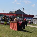 DSC_0301 Strażacy, którzy są dostępni do udziału w ćwiczeniach na trawiastym polu, gdzie znajduje się czerwony budynek i zbiornik wodny. W tle wydarzenia obserwowani widzowie oraz inni członkowie mojej rodziny.