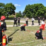 DSC_0308 Na obrazku widać grupę osób, w tym dzieci, ubranych w strażackie mundury uczestniczących w ćwiczeniach przeciwpożarowych na trawiastej przestrzeni. Walczący ich standard oraz widzowie, którzy robią zdjęcia i uczestniczą w doświadczeniach ćwiczeń.