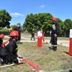 DSC_0312 Na obrazku strażacy w urządzeniach treningowych uczestniczyli w ćwiczeniach, obsługując węże i strumienie wodne na czerwono-białe, umieszczone na trawiastym polu. W tle widoczni są drzewa i grupa obserwatorów.