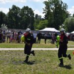 DSC_0347 Na obrazku widać dwa urządzenia biegnące przez trawiastą polanę i unoszące się węże strażackie podczas pokazu publicznego. W tle występują tłumy występujące oraz różne namioty, tworzące festynowe otoczenie.