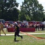 DSC_0348 Na obrazku przednim w czarnym mundurze ciągnie się na trawiastym polu, podczas gdy drugi ubrany na żółto. W tle są widoczne różne strażackie i tłum ludzi.