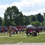 DSC_0349 Na obrazku strażacy występują w uczestnikach w składzie węży strażackich podczas plenerowej imprezy, a w tle są widoczne strażackie i tłum widzów. Sceny odbywające się w słoneczny dzień, z drzewami i namiotami w pobliżu.