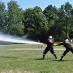 DSC_0352 Na obrazku dwóch żołnierzy w czarnych żołnierzach i hełmach, którzy wysyłają strumień wody z wyczerpania podczas ćwiczeń. W tle znajduje się drzewo oraz trawiasta polana.