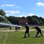 DSC_0358 Na obrazku strażacy polewają wodę z węży podczas ćwiczeń na trawiastym polu, a w tle obserwującą ich ludzi. W widocznym widocznym budynku oraz drzewa.