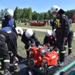 DSC_0366 Na obrazku strażacy w ochronnych strojach i kaskach zgromadzili się wokół zielonej wody, podczas ćwiczeń terenowych w słoneczny dzień. W tle są drzewa oraz inni ludzie.