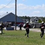 DSC_0373 Na obrazku strażacy w mundurze i hełmy biegną przez trawiasty teren podczas ćwiczeń dziennych. W tle znajdują się inne osoby w pobliżu budynku oraz sprzęt strażacki.