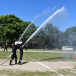 DSC_0383 Na obrazie dwóch żołnierzy w mundurach, którzy spryskują wodę z węży na zewnątrz podczas ćwiczeń treningowych w słoneczny dzień. W tle widoczni są inni strażacy oraz drzewa.