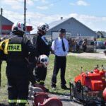 DSC_0389 Na obrazku strażacy w hełmach i mundurach gromadzą się wokół sprzętu gaśniczego rozłożonego na ziemi. Niedaleko stoi mężczyzna w koszuli i krawacie, a w tle są domy mieszkalne oraz kosze.