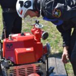 DSC_0391 Osoba wyposażona w urządzenia ochronne, które są objęte zamkniętą pompą strażacką na świeżym powietrzu, z połączonym wężem. W tle widoczni są obserwatorzy i widzowie.