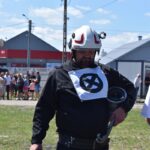 DSC_0393 Na zewnątrz znajduje się mężczyzna mający na sobie kask i kurtkę z innym symbolem, trzymający narzędzie w rękach. W tle, pod czystym niebem, zgromadzona jest grupa ludzi.