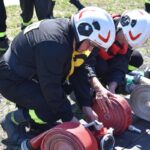 DSC_0394 Na obrazku dwóch funkcjonariuszy w kaskach i sprzęcie ochronnym, klęczących na ziemi i właściwychch zrolowanych przez funkcjonariuszy strażackich podczas treningu na świeżym powietrzu. Sceneria przygotowania do produkcji lub ćwiczeń doskonalących ich umiejętności.