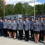 172-2654331 Na obrazku widać grupę umundurowanych funkcjonariuszy stojących w formacji na zewnątrz. W tle znajduje się drzewo oraz maszt z podniesionymi flagami.