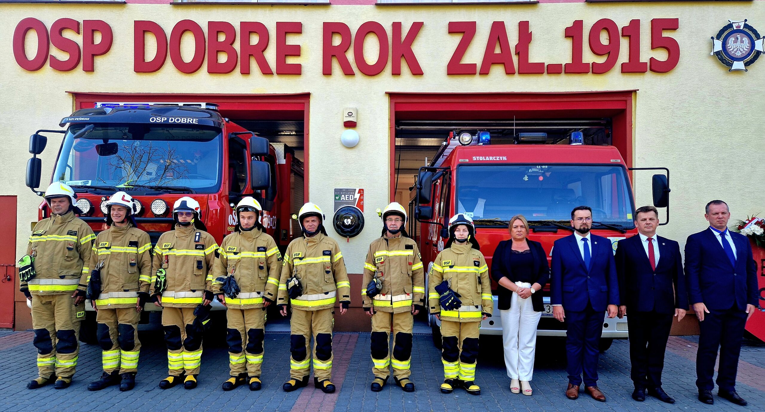 Na obrazku widać grupę ubranych w mundurach, którzy zostaną umieszczeni obok dwóch wozów strażackich. Po prawej stronie pięć osób ubranych formalnie, przed wprowadzeniem oznaczonym napisem „OSP Dobre Rok Zal.1915”.