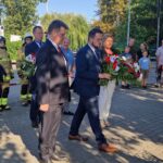 Na obrazku grupa ludzi, w tym mężczyznach w garniturach i kobieta ubrana na biało, składane wieńce kwiatowe pod pomnikiem. Obok znajduje się strażacy i obserwatorzy kierujący ceremonii.