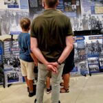 Na obrazku trzy osoby blisko siebie, oglądając wystawę z historycznymi fotografiami i panelami tekstowymi. Cała scena ma miejsce w dobrze oświetlonym utworze.